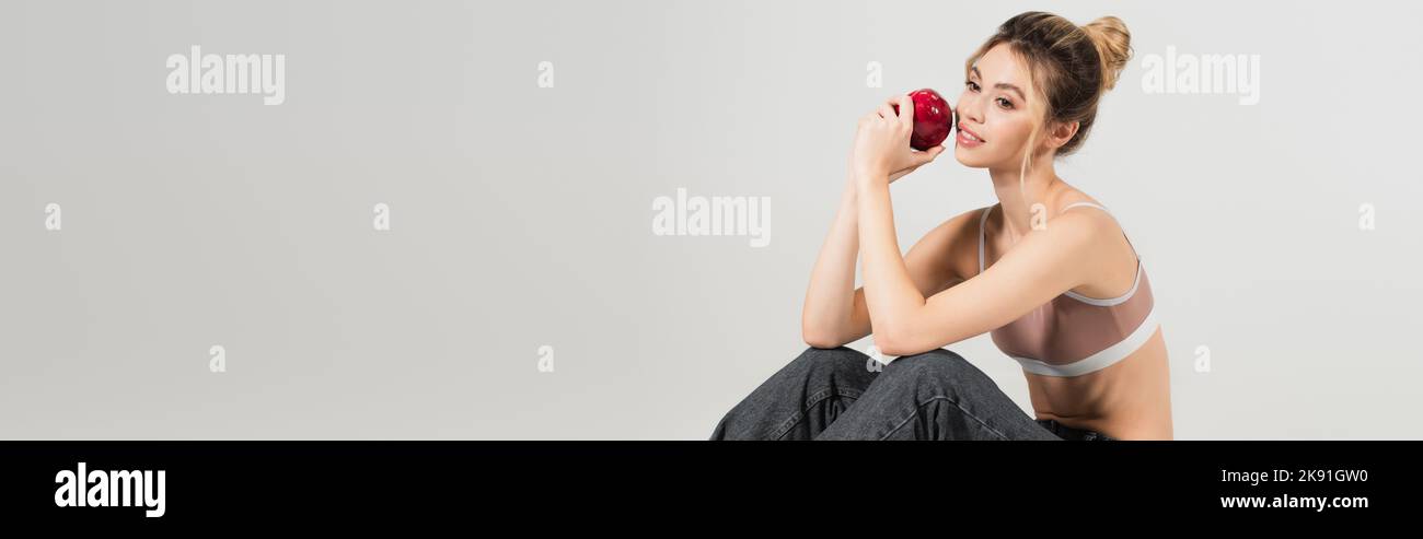 Positive junge Frau in Sport-Top sitzen mit reifen Apfel isoliert auf grau, Banner Stockfoto