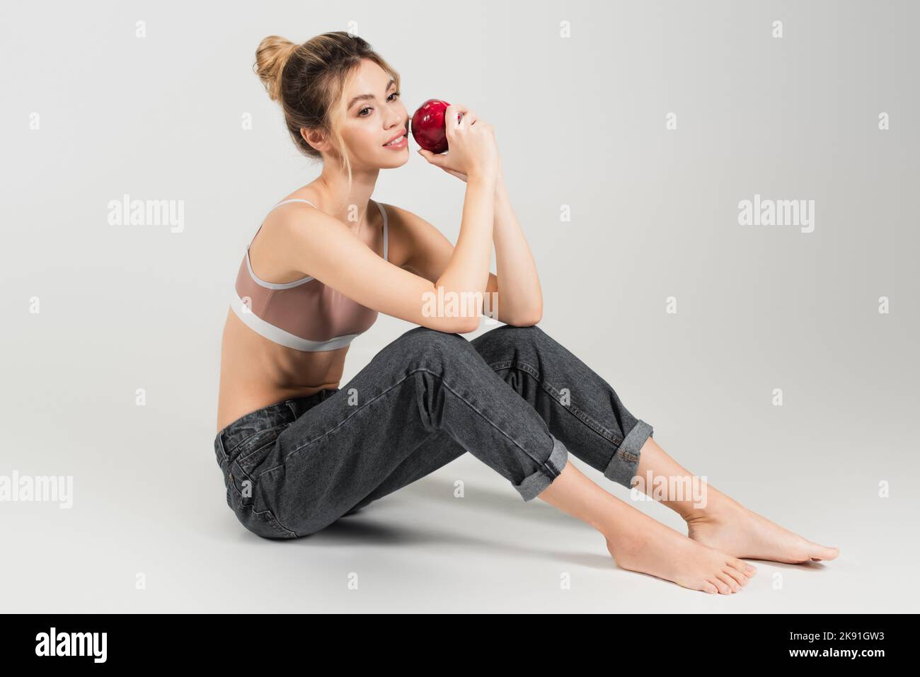 Junge Barfußfrau mit perfektem Körper und Haut, die mit frischem Apfel auf grauem Hintergrund sitzt Stockfoto