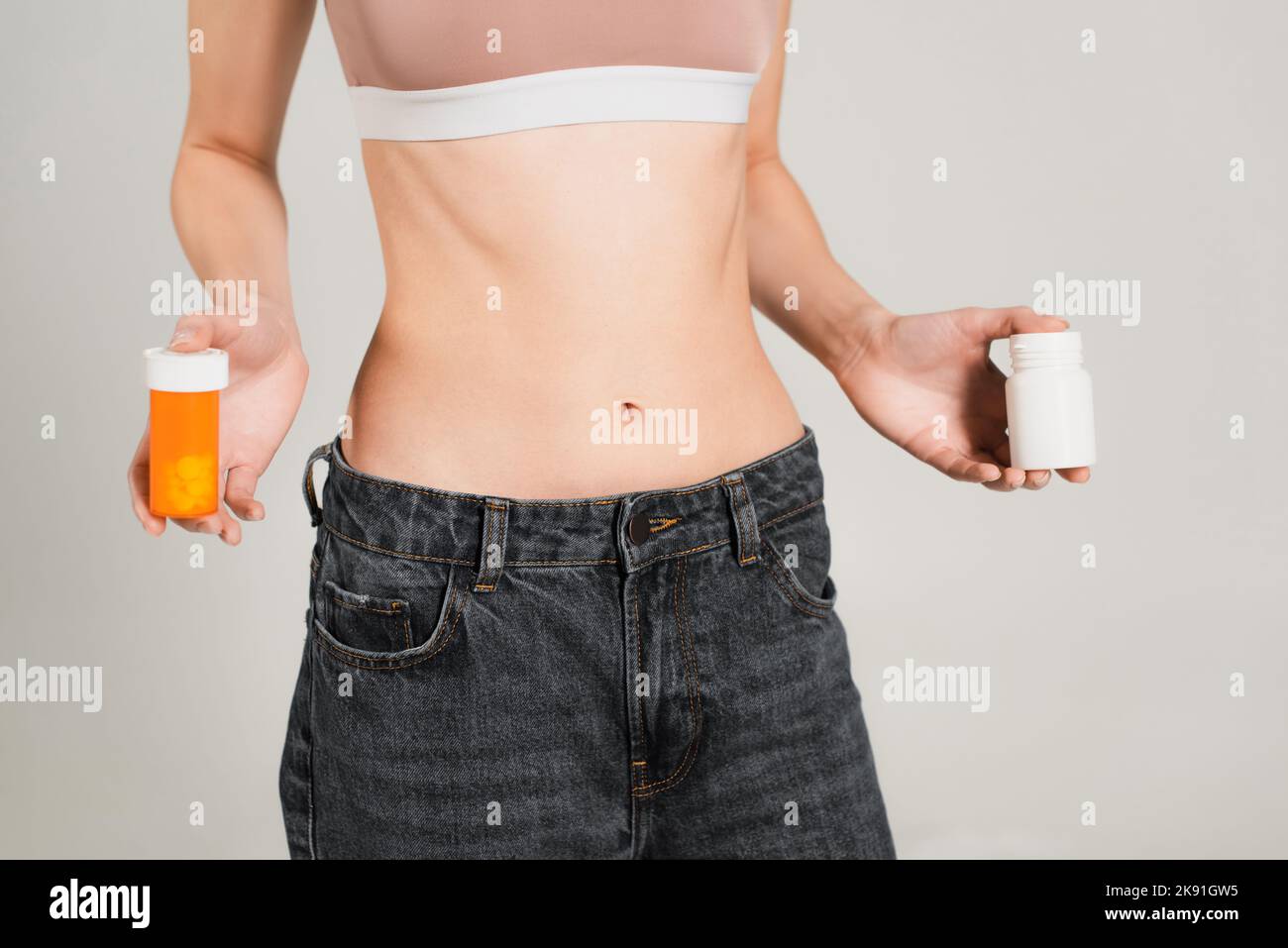 Teilansicht einer schlanken Frau in Jeans, die Behälter mit Vitaminen auf Grau isoliert hält Stockfoto