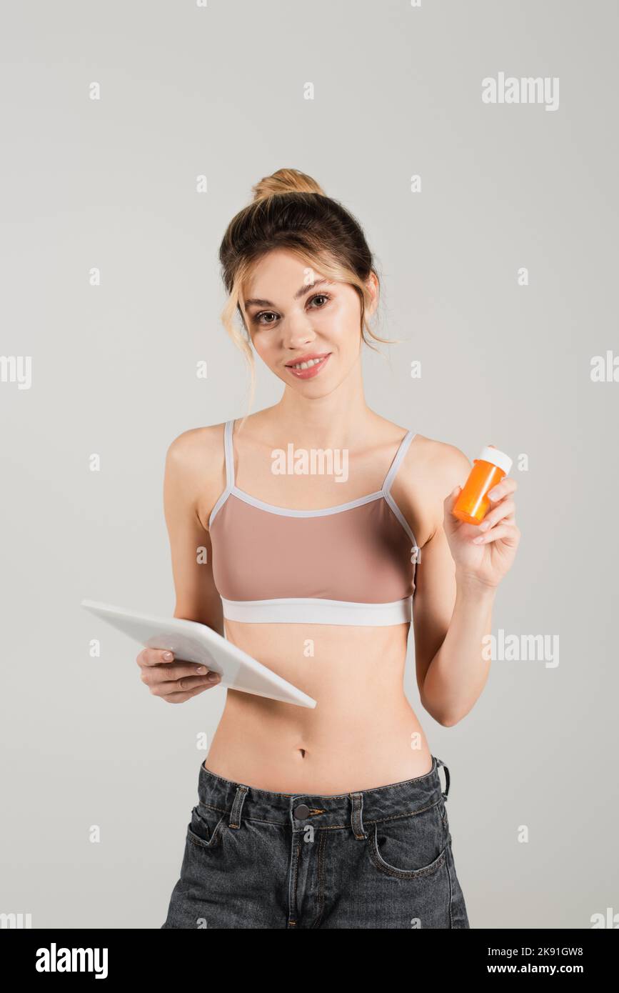 Fit und gesunde Frau in Sport-BH hält digitale Tablette und Vitamine isoliert auf grau Stockfoto