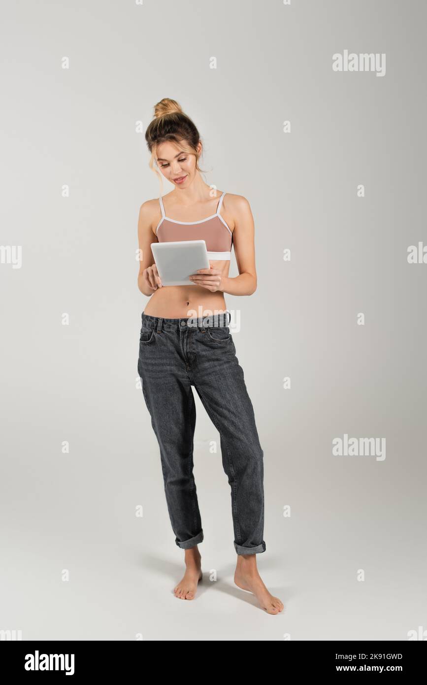 Die Frau in voller Länge sitzt barfuß in Jeans und Sporttop mit digitalem Tablet auf grauem Hintergrund Stockfoto