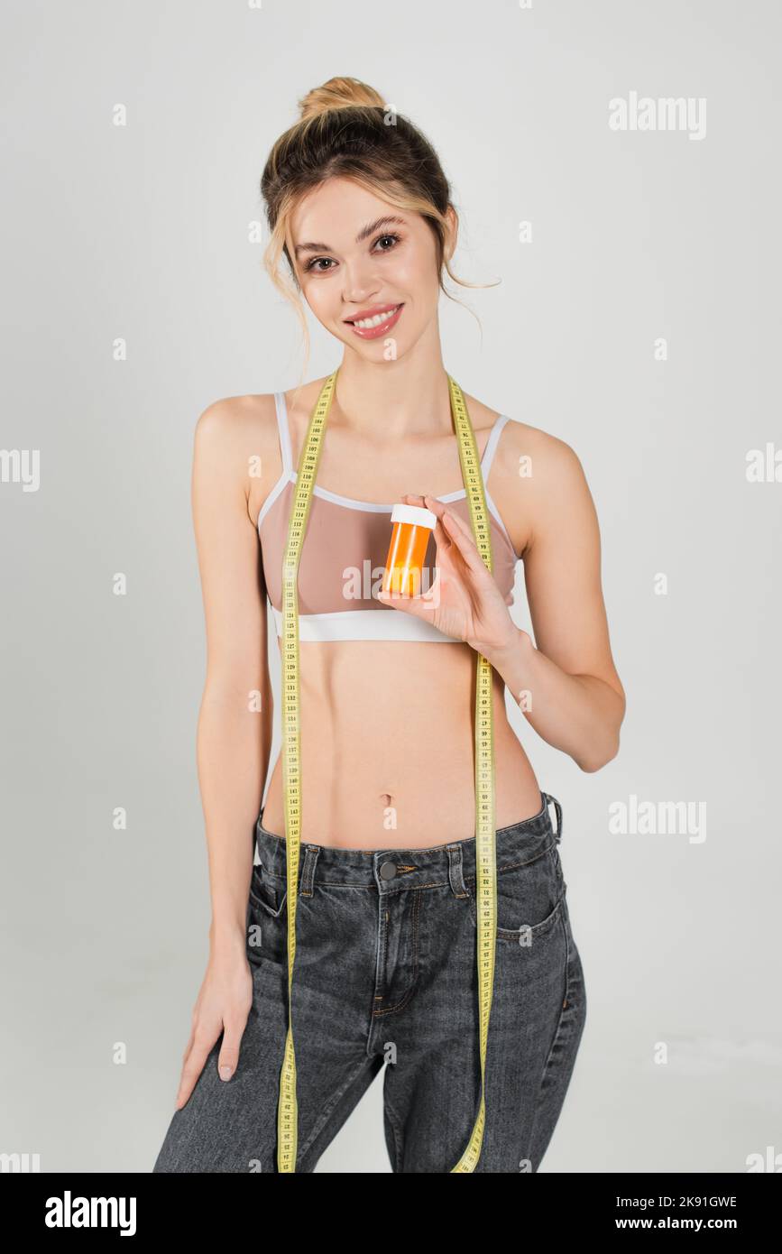 Junge Frau mit schlanker Figur stehend mit Maßband und Vitaminen isoliert auf grau Stockfoto