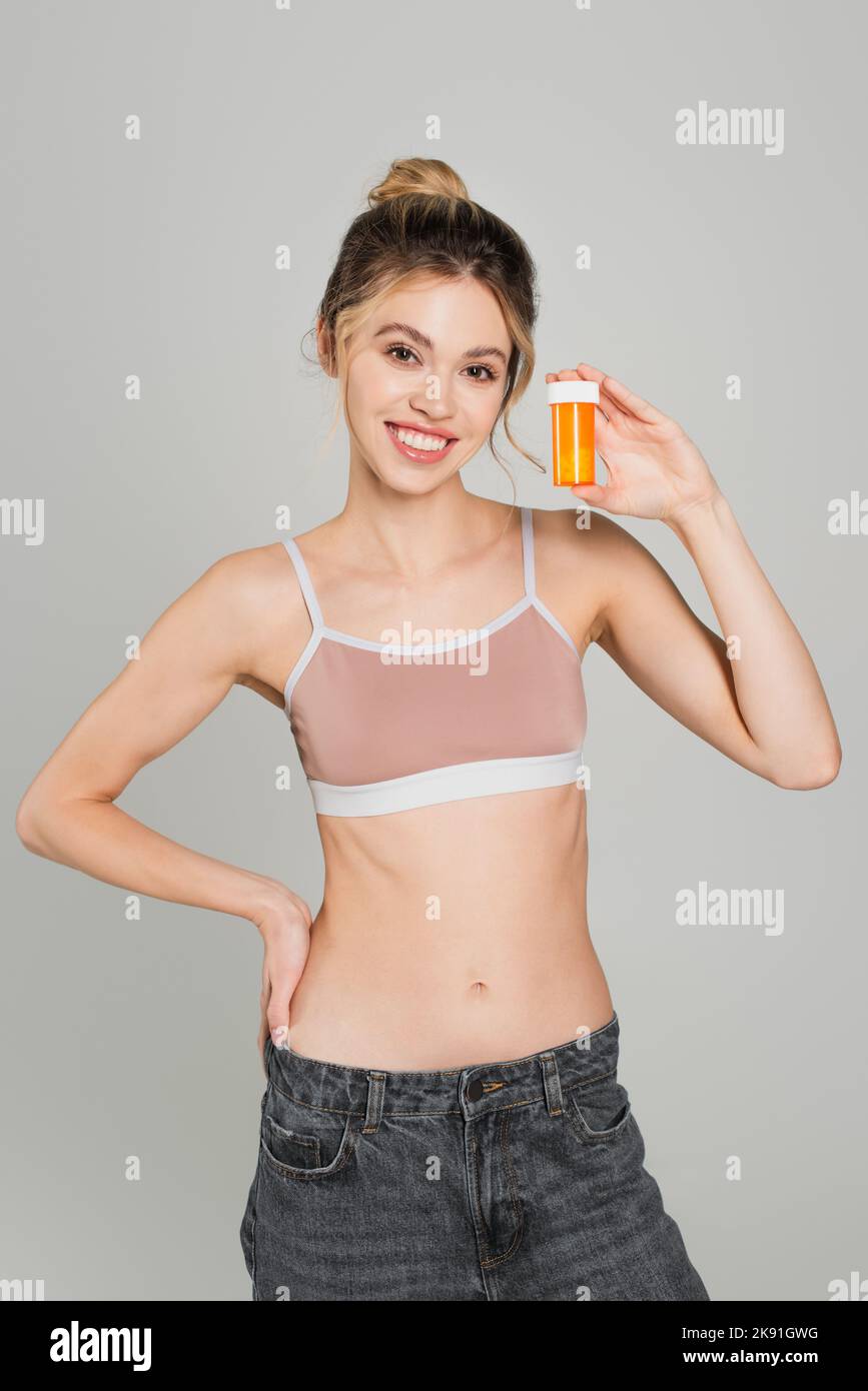 Fröhliche und fit Frau in Sport-Top mit Hand auf der Hüfte und zeigt Behälter mit Vitaminen isoliert auf grau Stockfoto