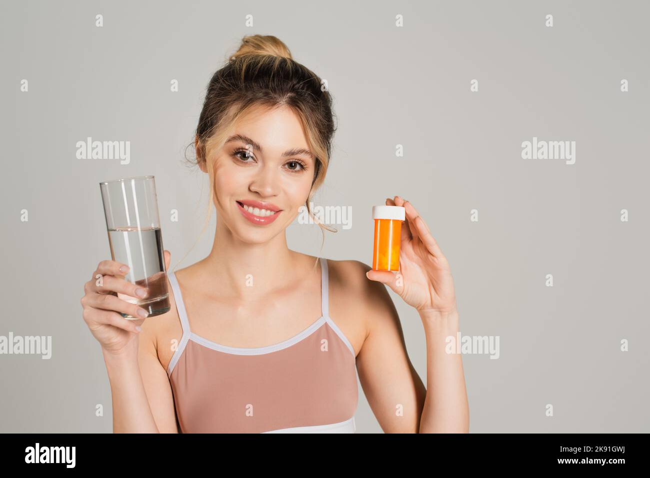 Frau mit perfekter Haut lächelt der Kamera zu, während sie ein Glas Wasser und Vitamine auf Grau isoliert hält Stockfoto