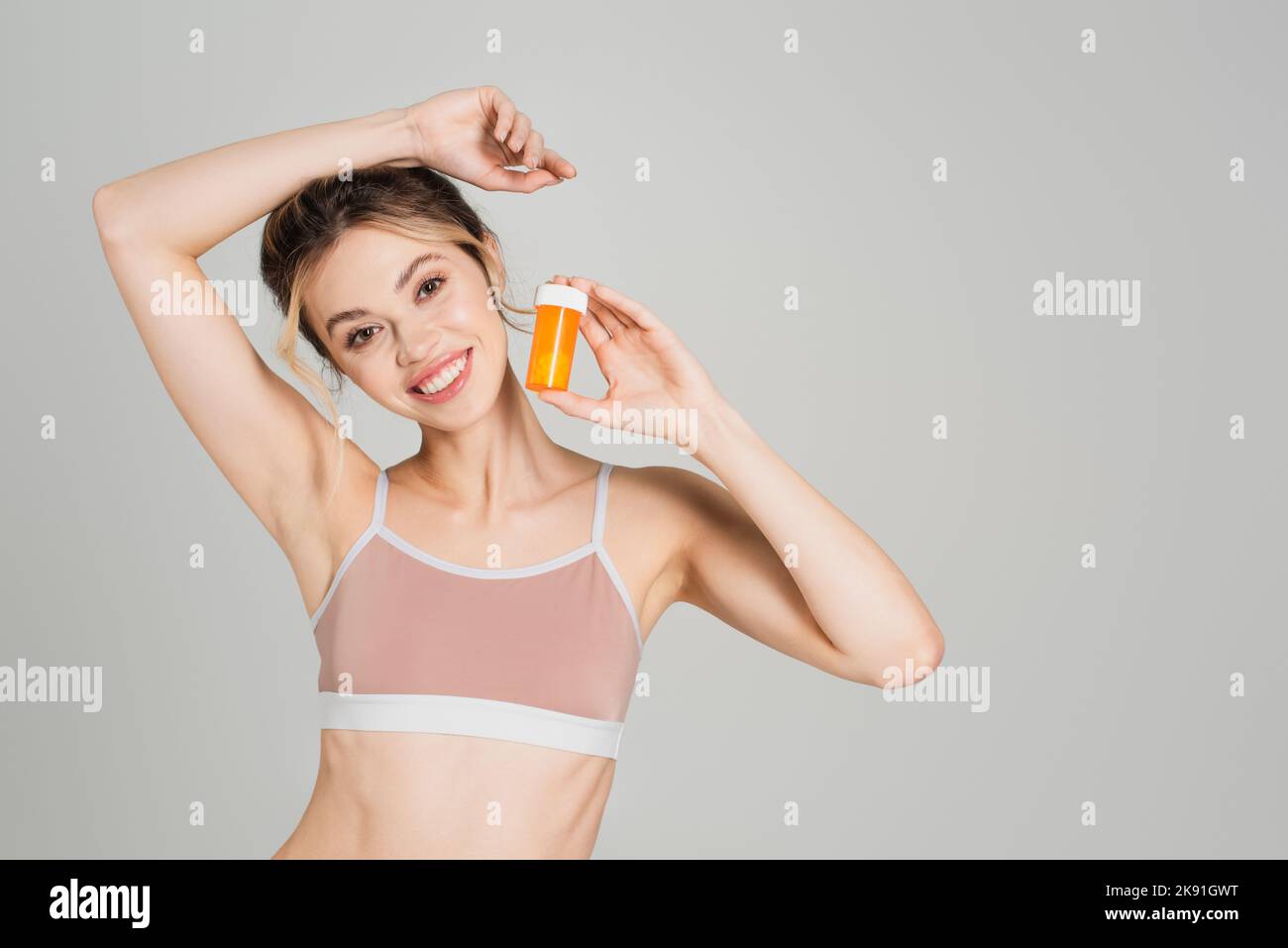 Aufgeregt und schlanke Frau mit natürlichem Make-up hält Vitamine und lächelt auf Kamera isoliert auf grau Stockfoto