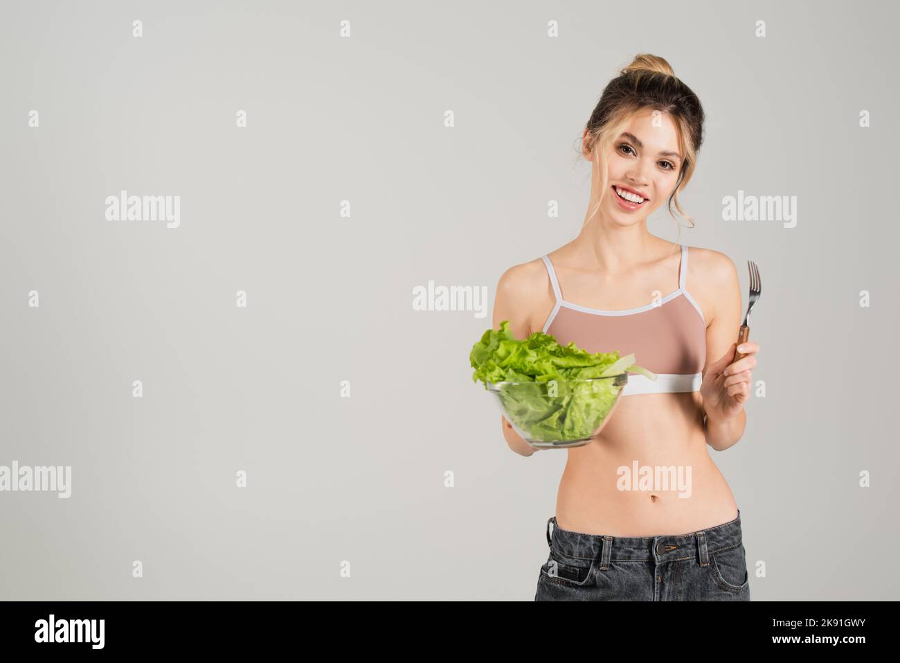 Junge Frau mit schlankem Körper, der Gabel und frischen Salat hält, während sie auf grau isoliert auf die Kamera lächelt Stockfoto