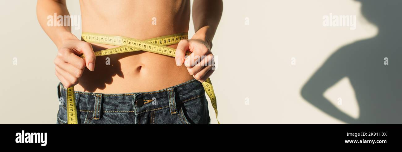 Teilansicht einer schlanken Frau in Jeans, die die Taille auf weißem Hintergrund mit Schatten und Banner misst Stockfoto