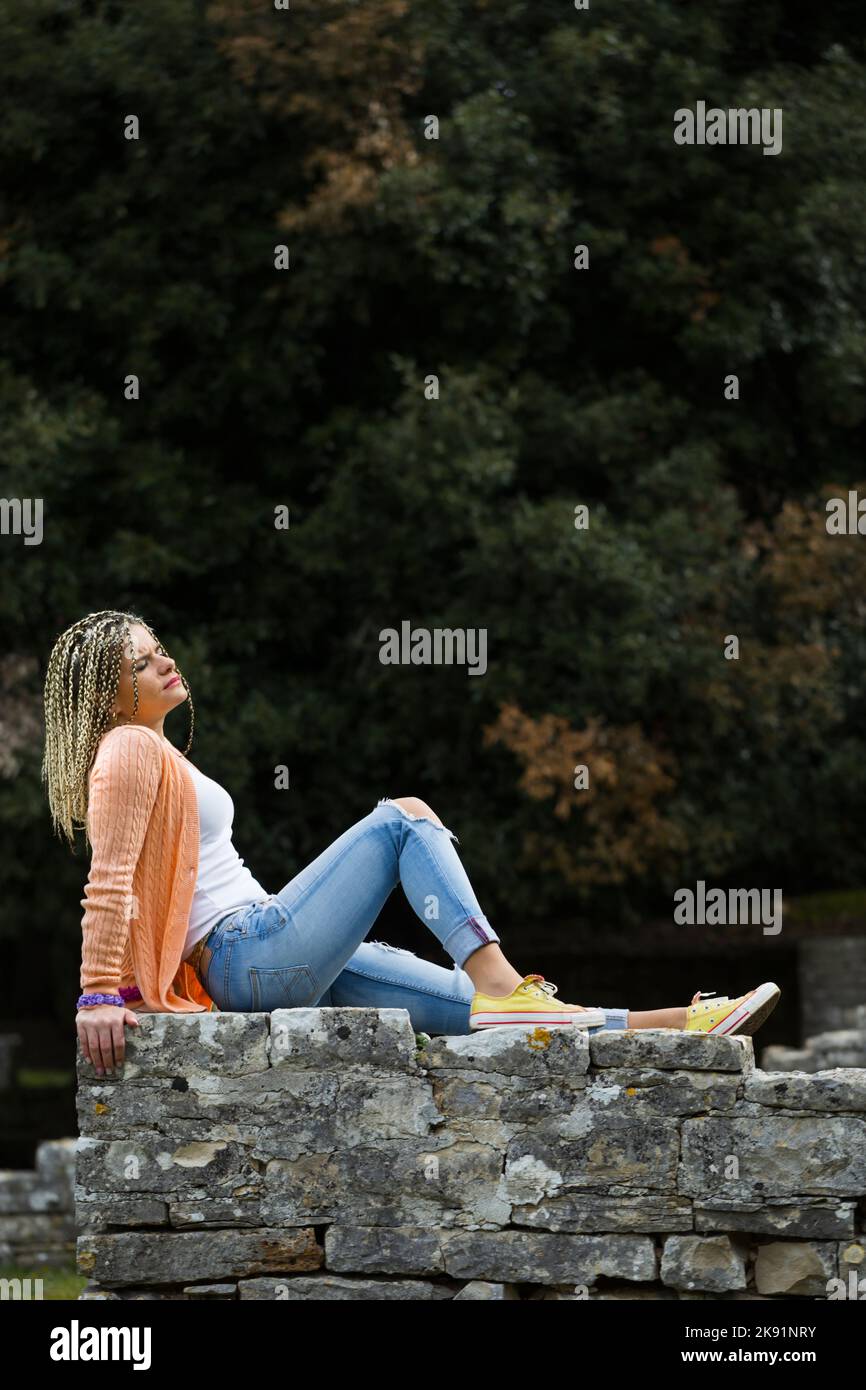 Attraktive reife Frau mit blonden Haaren aus fairhair und lässiger Kleidung, die auf einer Steinwand sitzend, zurückliegend und zurückgelehnt posiert Stockfoto