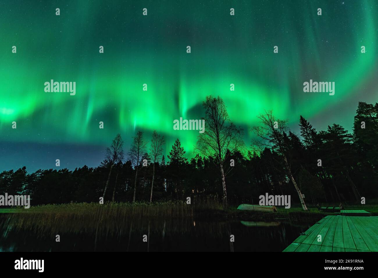 Atemberaubende, kräftige grüne Lichter der tanzenden Aurora über Nordwald, See, kleinem Boot, Brücke. Stocksjo See, Nordschweden, Umea Stadt, Nacht Stockfoto