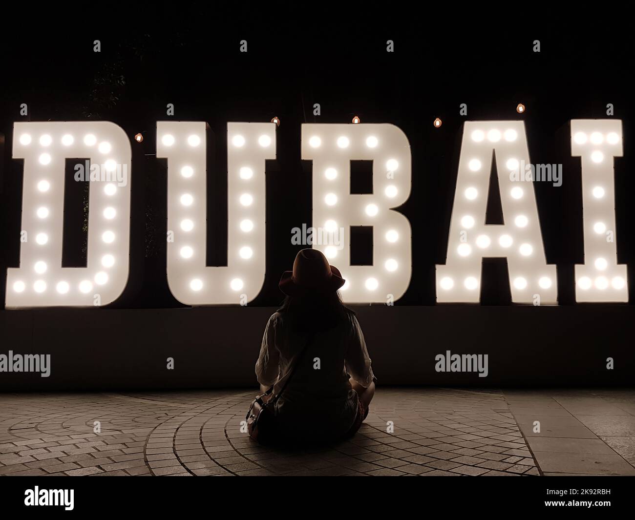 Frau sitzt mit gekreuzten Beinen auf dem Boden, vor dem Hintergrund des großen Dubai-Festzelttextes. Frau schaut auf Briefe, zurück in Richtung Kamera, in der Nähe Silhouette in der Nacht. Stockfoto