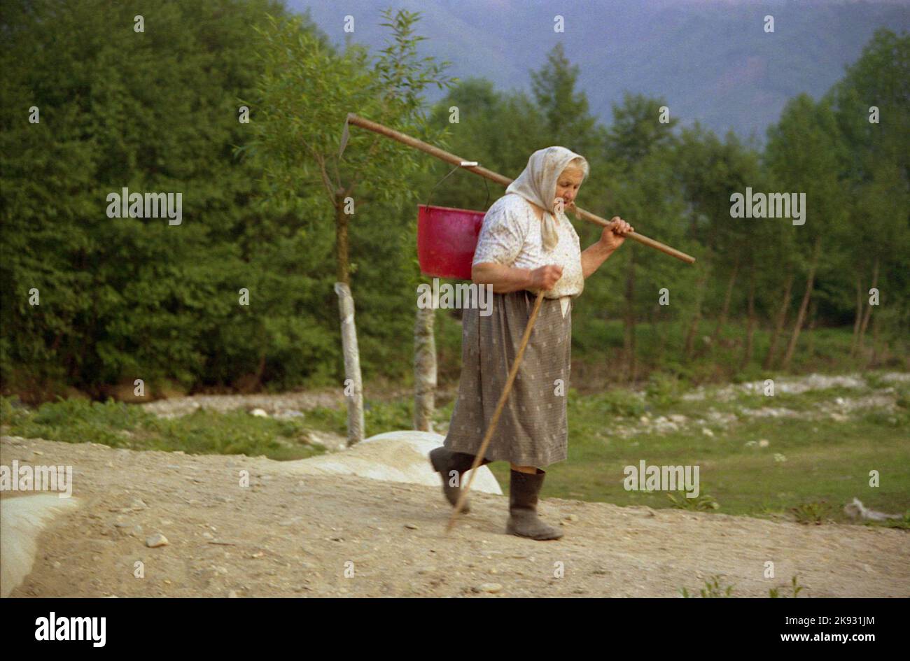 Covasna County, Rumänien, ca. 2002. Ältere Frau auf dem Land, die mit einem Stock geht und einen schweren Eimer trägt. Stockfoto