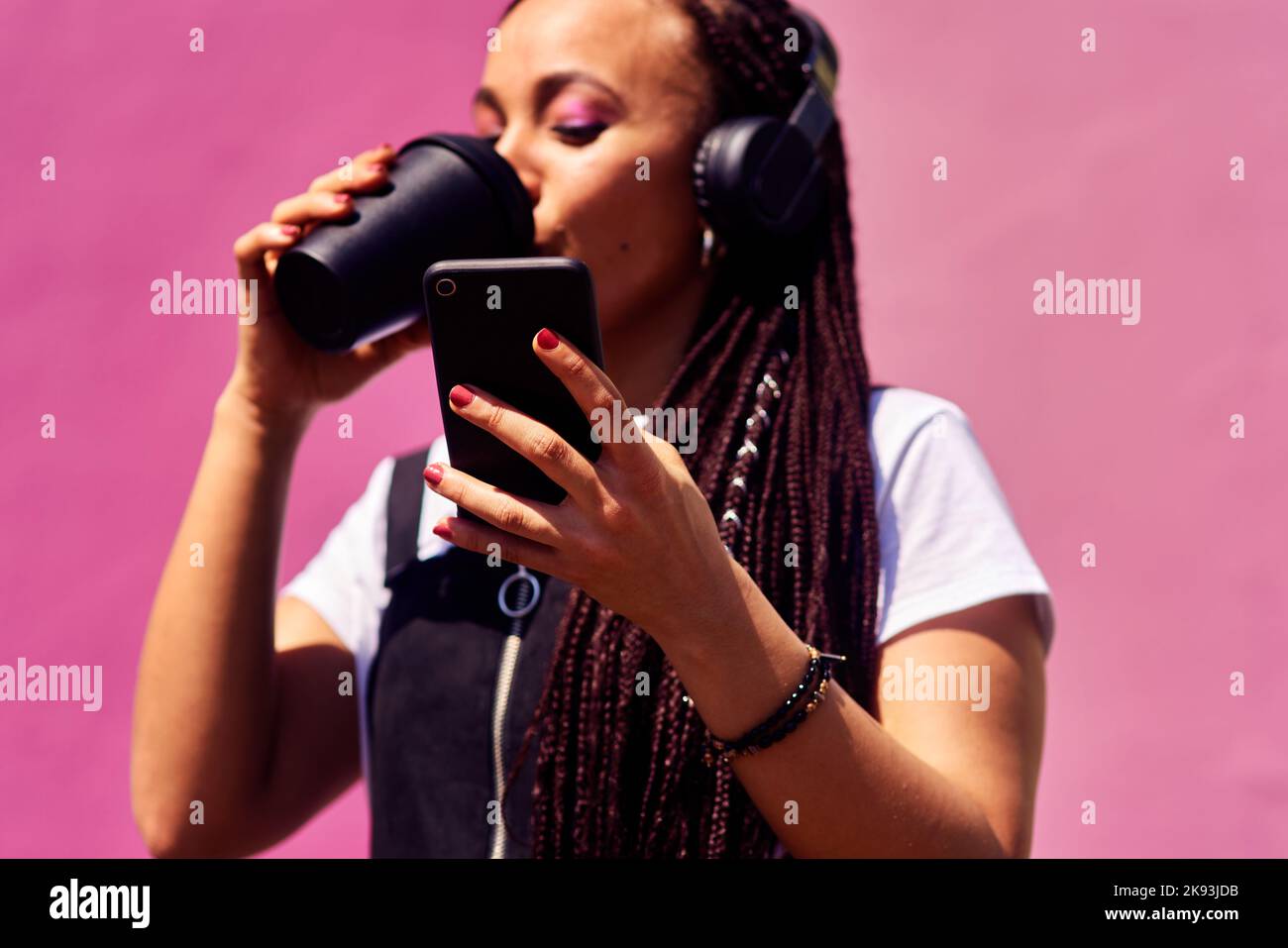 Kaffee und Technik sind Millennials Notwendigkeiten. Eine attraktive junge Frau, die allein gegen eine rosa Wand steht und Kaffee trinkt, während sie Technologie benutzt. Stockfoto