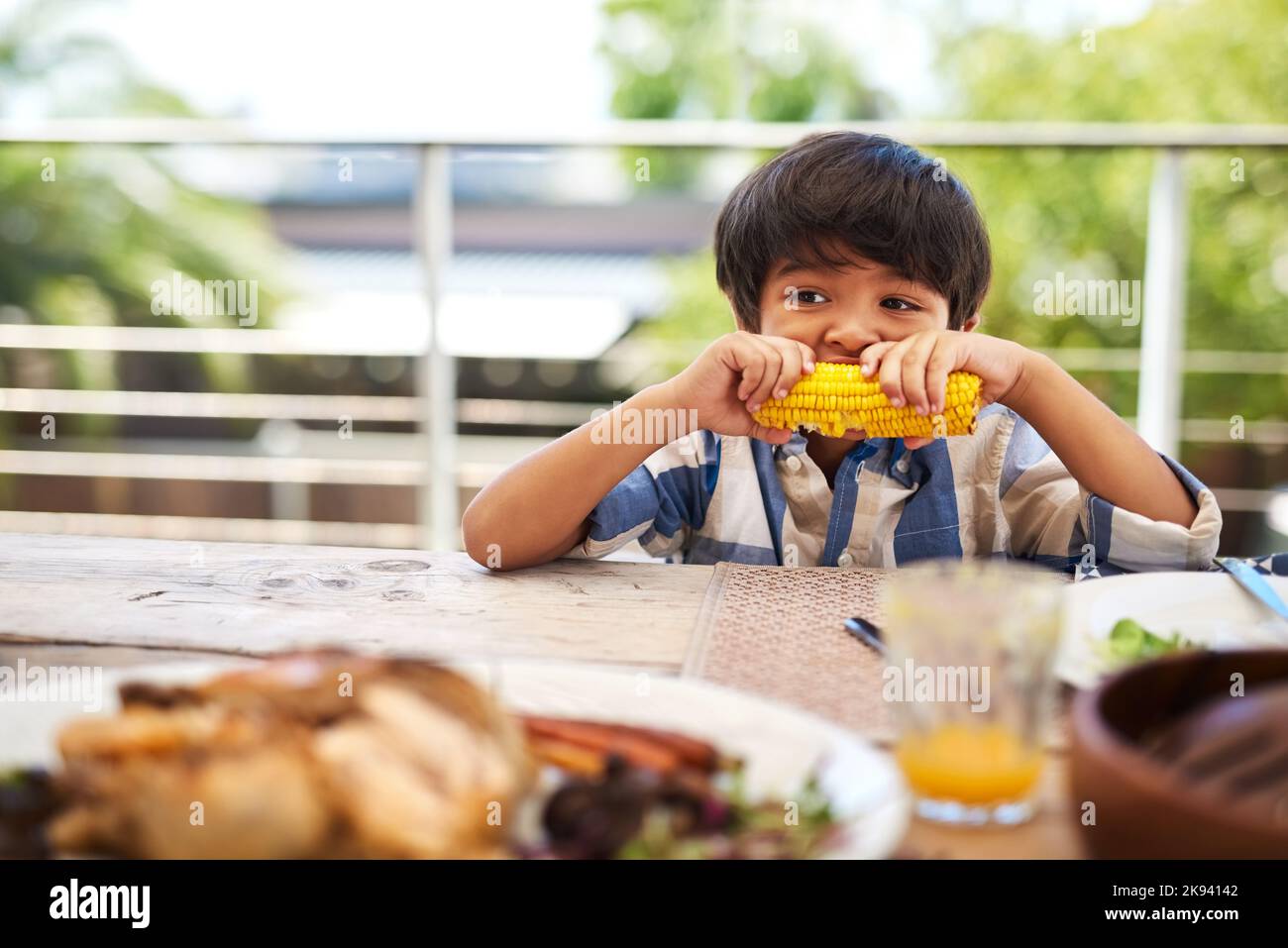 Nichts schmeckt besser als ein gelbes Gemüse. Ein entzückender kleiner Junge, der draußen an einem Tisch eine Maiskolben isst. Stockfoto