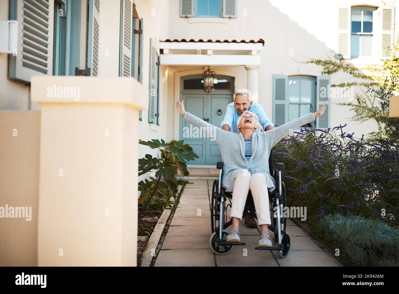 Lebten noch ein Leben voller Nervenkitzel. In voller Länge ein fröhlicher älterer Mann, der seine Frau zu Hause in einem Rollstuhl in ihrem Hinterhof schiebt. Stockfoto
