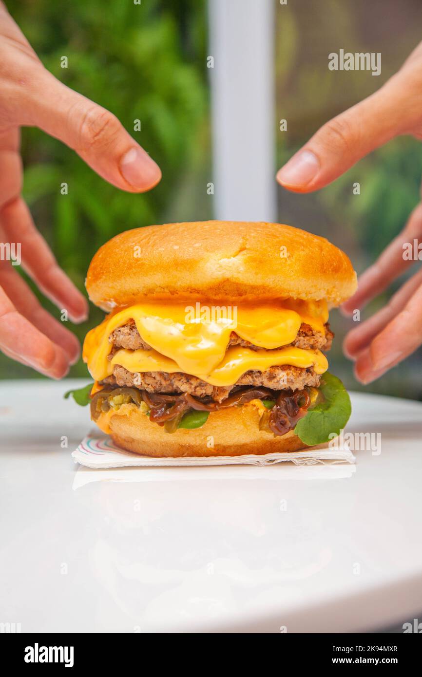 Hände im Begriff, einen Queso geschmolzenen Käseburger mit üppigem grünem Hintergrund in einem Café zu heben. Stockfoto
