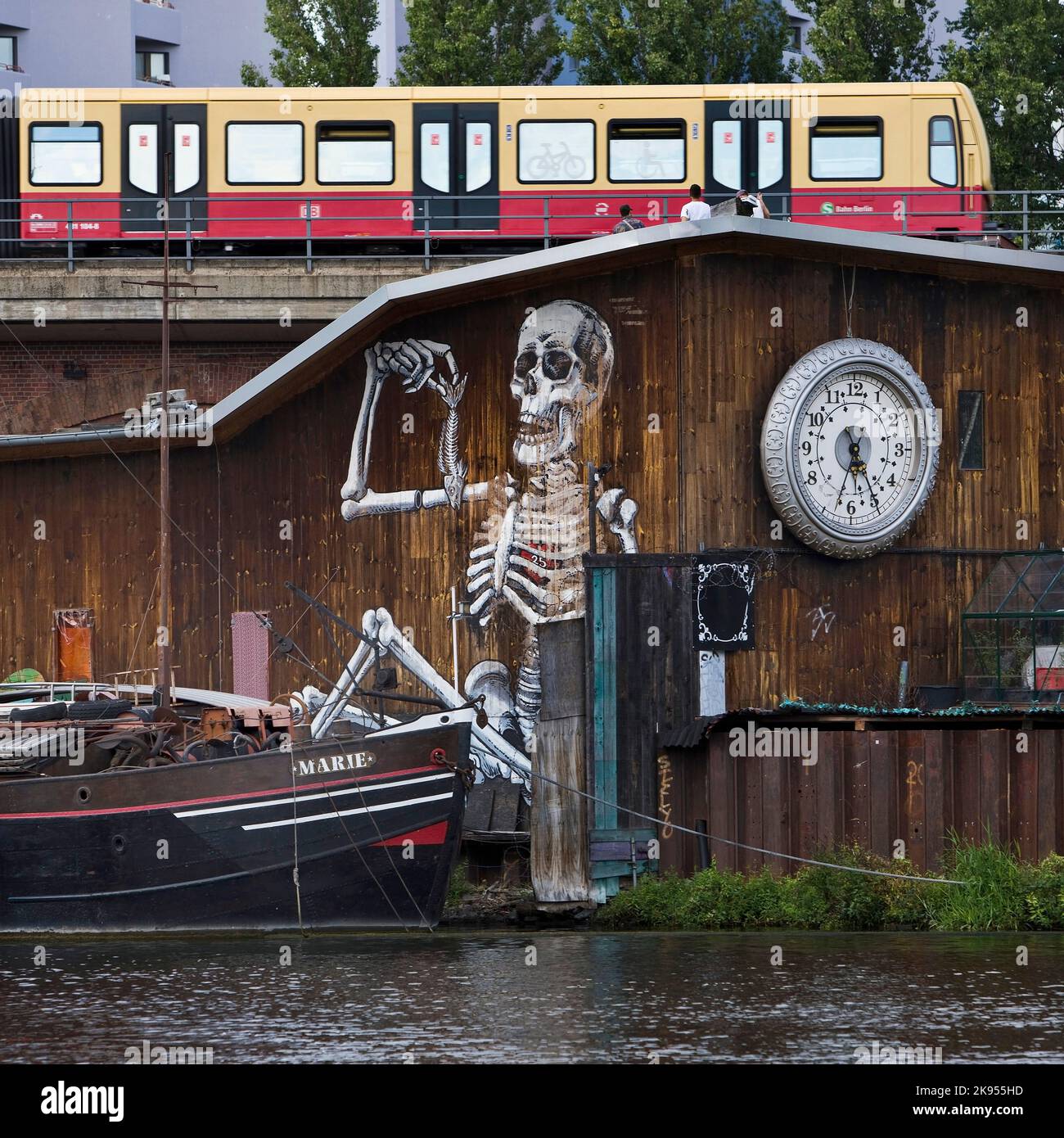 Holzwand mit Uhr und bemaltem Skelett auf der Spree mit Schiff und fahrender S-Bahn bei der Michaelbrücke, Deutschland, Friedrichshain, Berlin Stockfoto