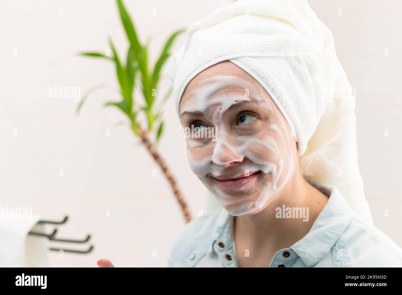 Eine junge, schöne Frau mit einem weißen Handtuch auf dem Kopf im Badezimmer brachte Make-up-Entferner vor dem Hintergrund einer grünen Pflanze auf ihr Gesicht. Sel Stockfoto