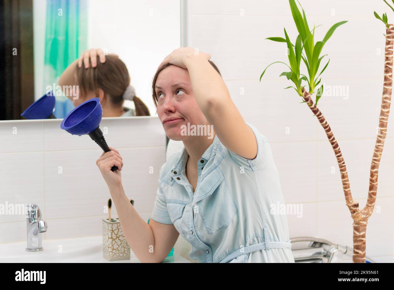 Eine junge Putzfrau in einem hellen Badezimmer mit einem Kolben, um die Verstopfung in ihren Händen zu beseitigen. An der Wand befindet sich ein Spiegel mit Spiegelung. Grüne Pflanze. Stockfoto