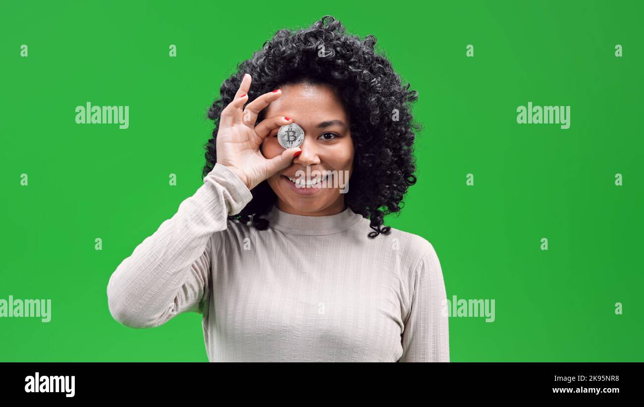 Eine isolierte afroamerikanische Frau auf einem grünen Bildschirm lächelt und hält einen versilberten Bitcoin vor ihrem Auge Stockfoto