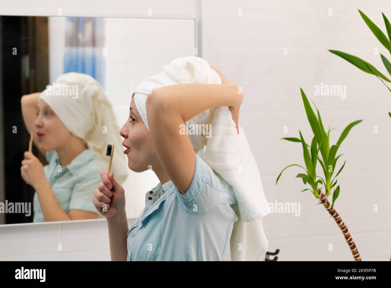 Eine junge hübsche Frau in einem blauen Bademantel mit einem Handtuch auf dem Kopf in einem hellen Badezimmer mit einer Zahnbürste in der Hand singt Lieder. Reflexion im mir Stockfoto