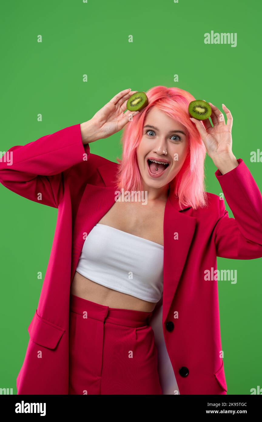 Fröhliche junge Frau mit Kiwifruit, die an der Wand steht Stockfoto