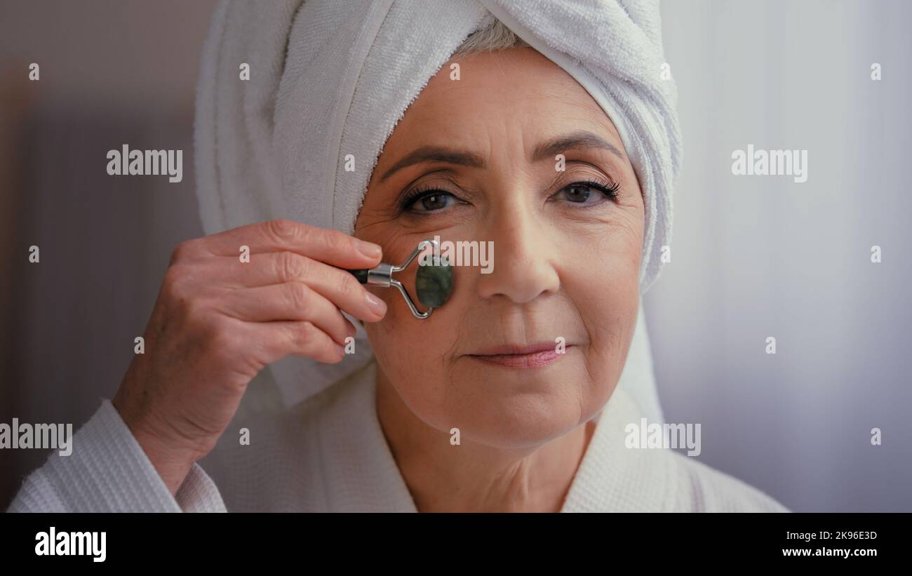 60s ältere ältere kaukasische Frau mittleren Alters mit Handtuch auf dem Kopf im Bad morgendliche Gesichtsmassage mit Steinwalze Ältere schöne Dame Hautpflege Stockfoto