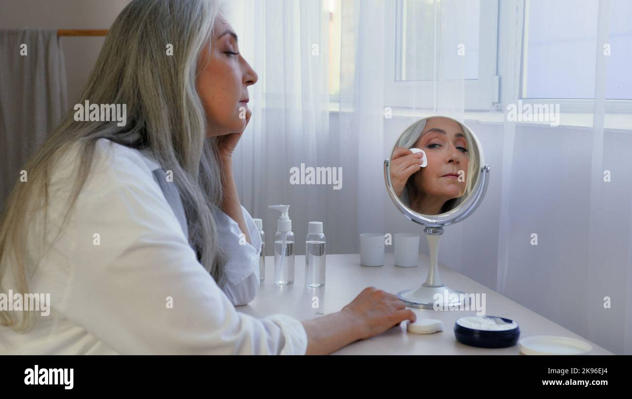 Grauhaarige kaukasische reife Dame mittleren Alters weiblich alt 60s Frau zu Hause suchen Spiegel wischt Gesicht mit Wattepad mit Tonic Lotion Serum Abstriche Stockfoto