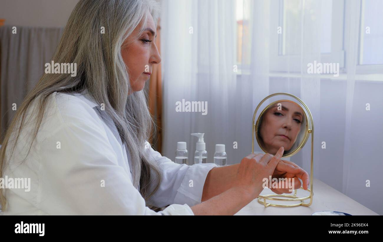 Alte ältere grauhaarige Frau am Morgen im Schlafzimmer Schönheitsverfahren Gesundheit Hautpflege. 60s reife Frau mittleren Alters, die sich mit Falten die Hände rieb Stockfoto