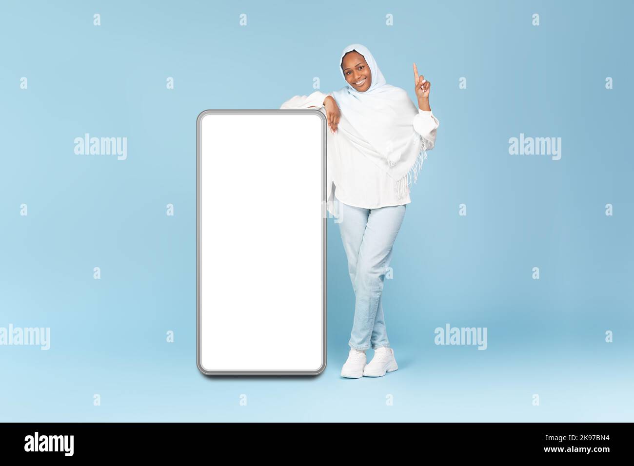 Werbung für mobile Anwendungen. Die ganze Körperlänge einer schwarzen muslimischen Dame zeigt den Finger nach oben und lehnt sich auf ein großes Smartphone mit leerem Bildschirm Stockfoto