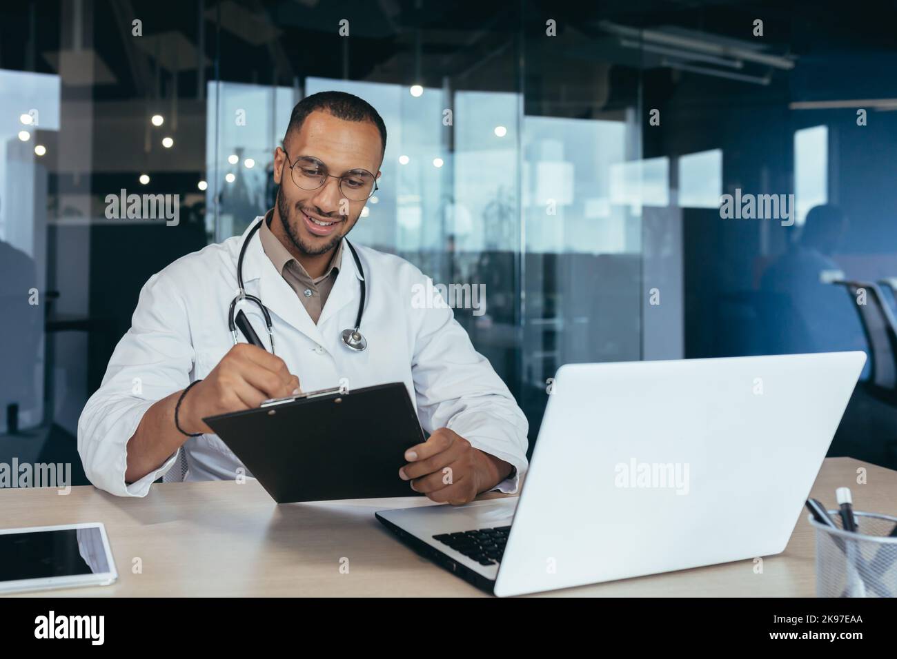 Online-Empfang im Krankenhaus. Junge gutaussehende hispanische Ärztin, die über einen Laptop mit dem Patienten spricht. Er sitzt im Büro am Arbeitstisch, schreibt die Geschichte des Hustens, die Beschwerden des Patienten und das Rezept für Medikamente auf. Stockfoto