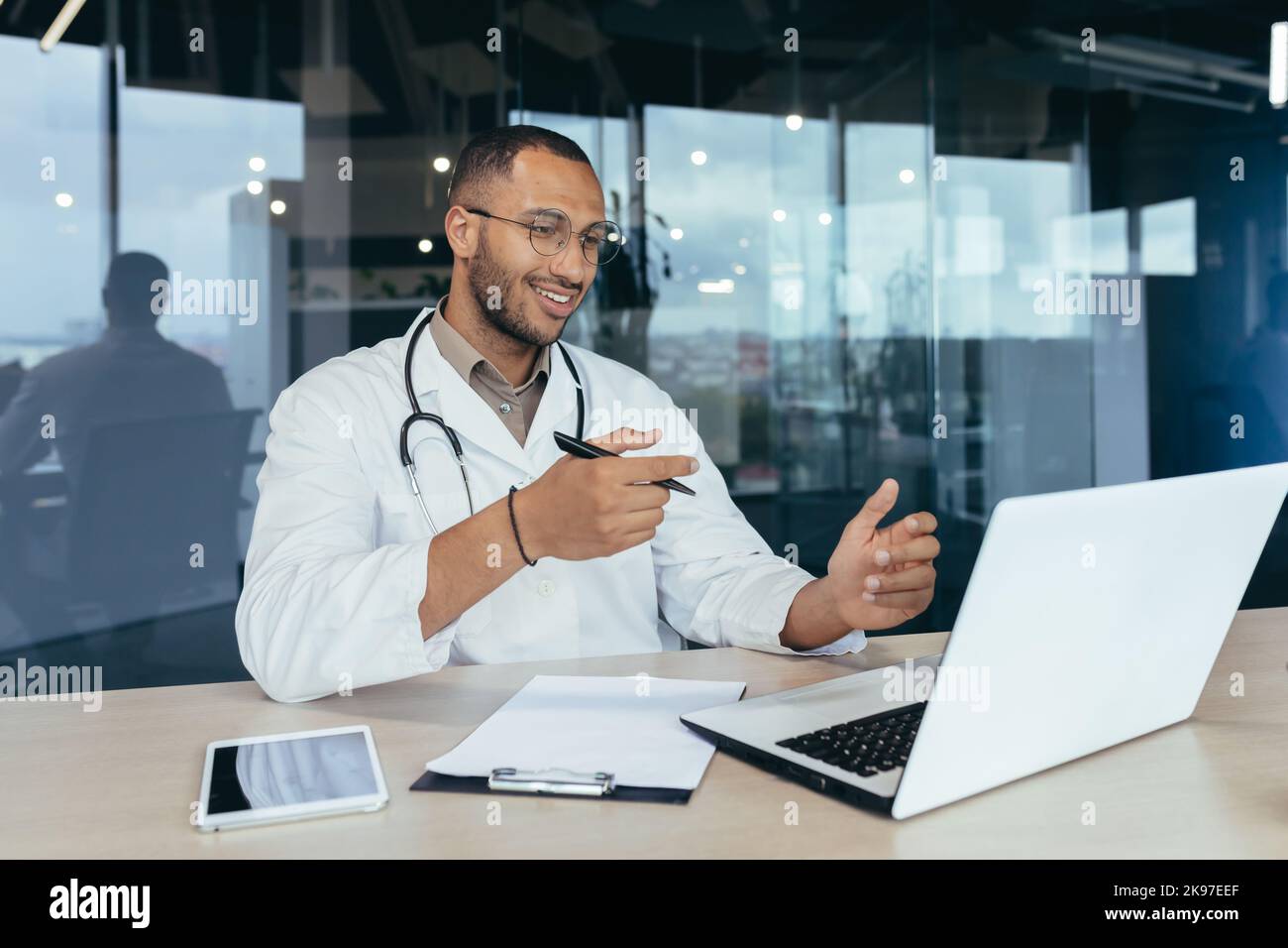 Online-Empfang im Krankenhaus. Junger gutaussehender hispanischer Arzt, der mit einem Patienten über einen Videoanruf von einem Laptop spricht. Sitzt im Büro am Schreibtisch, spricht mit der Kamera, erklärt und winkt ihm die Hände Stockfoto