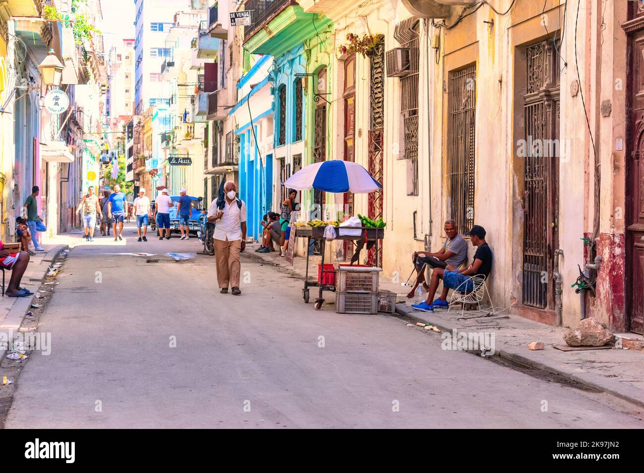 Zwei Männer verkaufen Obst und Gemüse in einem Wagen. Verwitterte Häuser stehen auf der Straße. Andere Kubaner sind in der Nachbarschaft auf dem Vorweg. Stockfoto