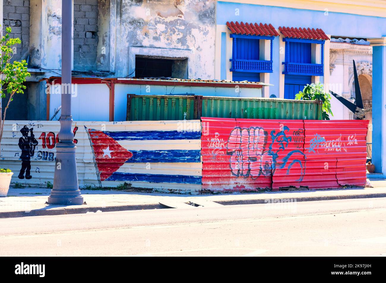 Ein Frachtcontainer ist von einem metallischen Zaun mit Graffiti und einer kubanischen Flagge umgeben. Im Hintergrund alte Gebäude. Stockfoto