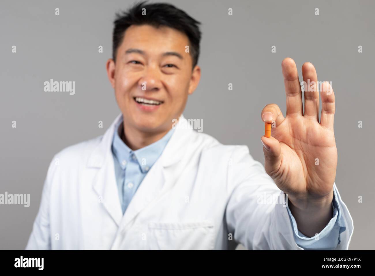 Lächelnder koreanischer Arzt mittleren Alters in weißem Mantel zeigt auf grauem Hintergrund isolierte Medikamentenkapsel Stockfoto