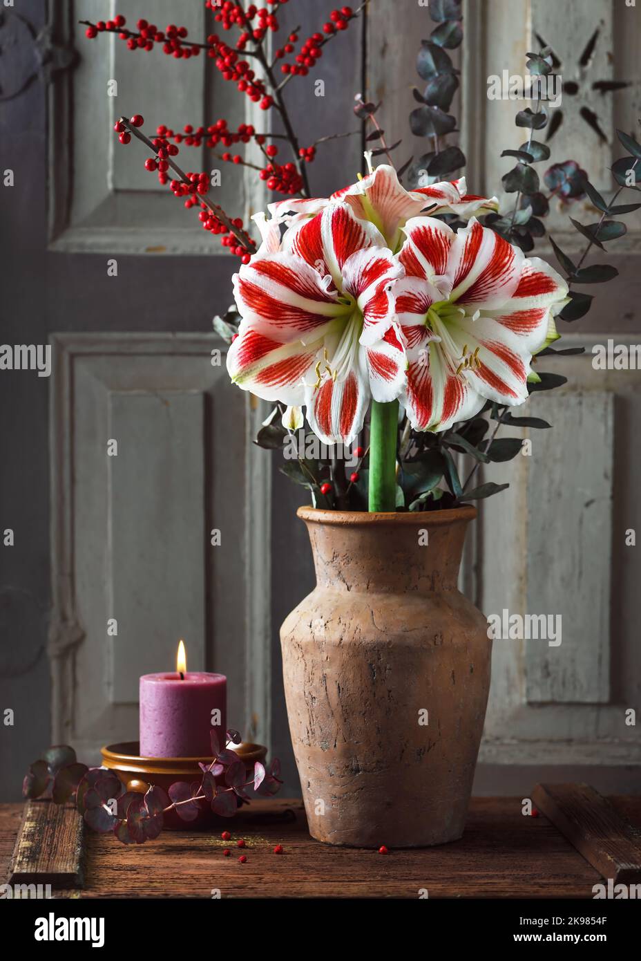 Bouquet mit roten, weißen Amaryllis-Blüten, Eukalyptus und roten Stechbeerzweigen, in einer Keramikvase und brennender Kerze in einer kleinen Schüssel. Stockfoto