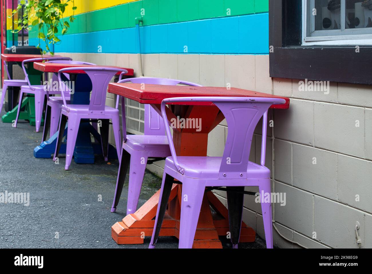 Ein Restaurant oder Cafe im Freien mit hellgelben, grünen und blauen Strichen an der Außenwand. Die Terrassentische sind farbenfroh aus rotem Metall Stockfoto