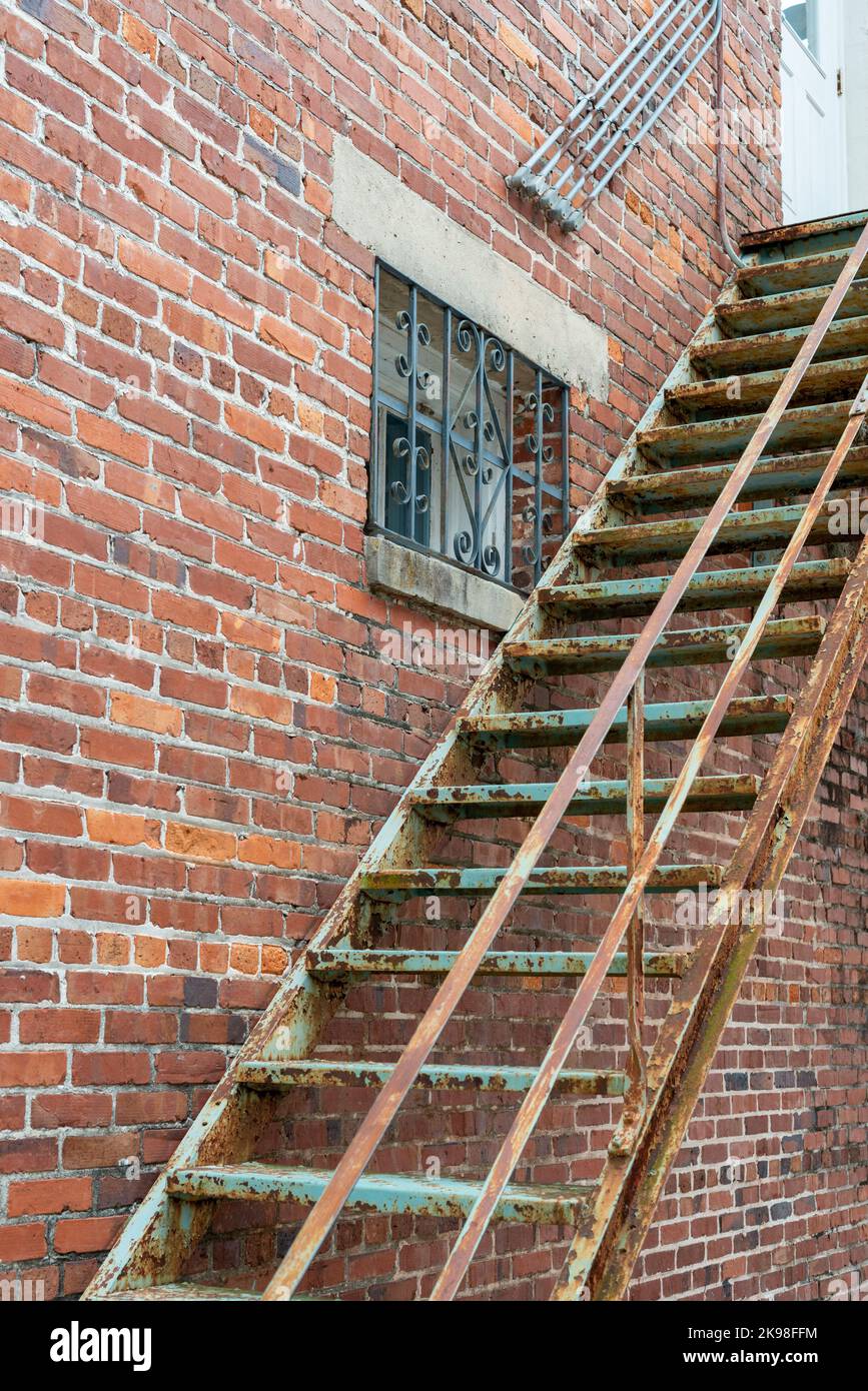 Die Eckmauer eines alten braunen und roten Ziegelgebäudes mit rostigen Metall-Feuertreppen. Es gibt ein Fenster mit mehreren Fenstern Stockfoto