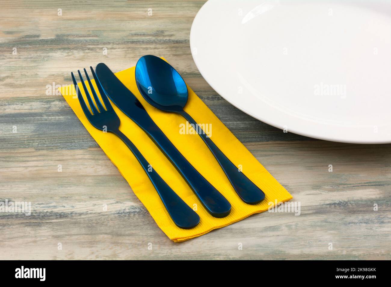 Blaues Gabelmesser und Löffel auf gelber Serviette neben weißem Teller Stockfoto
