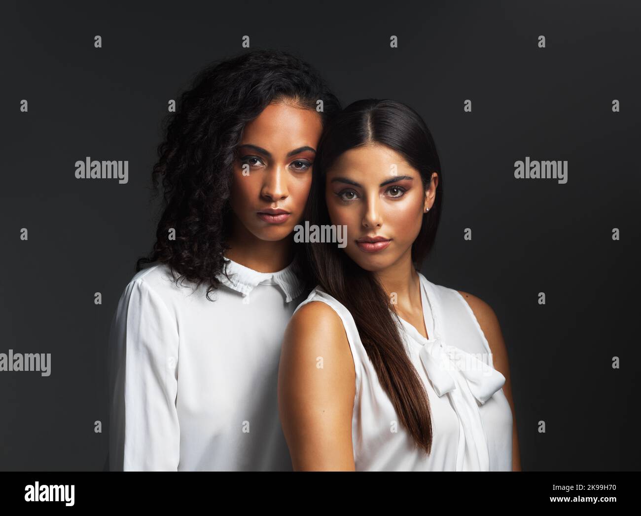Sieht wild aus. Porträt zweier attraktiver junger Frauen in weißen Blusen, die eng zusammen vor dunklem Hintergrund im Studio posieren. Stockfoto