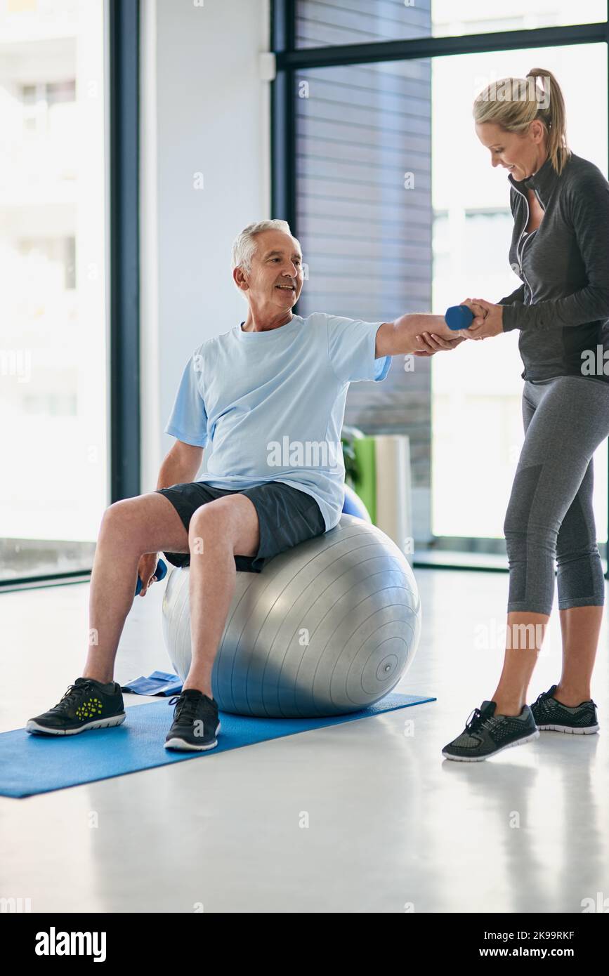 Lassen Sie uns versuchen, diese für ein paar Sekunden zu halten. Ganzkörperaufnahme eines glücklichen älteren Mannes, der mit Hilfe eines Physiotherapeuten mit Gewichten arbeitet. Stockfoto