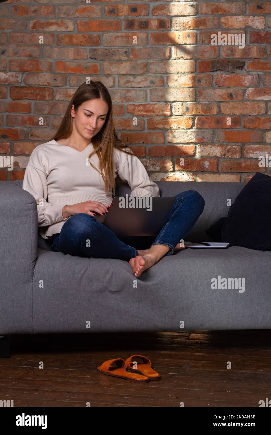 Frau in einem weißen Pullover, die mit einem Laptop arbeitet, mit einem Notebook an der Seite und Schuhen vor ihr Stockfoto