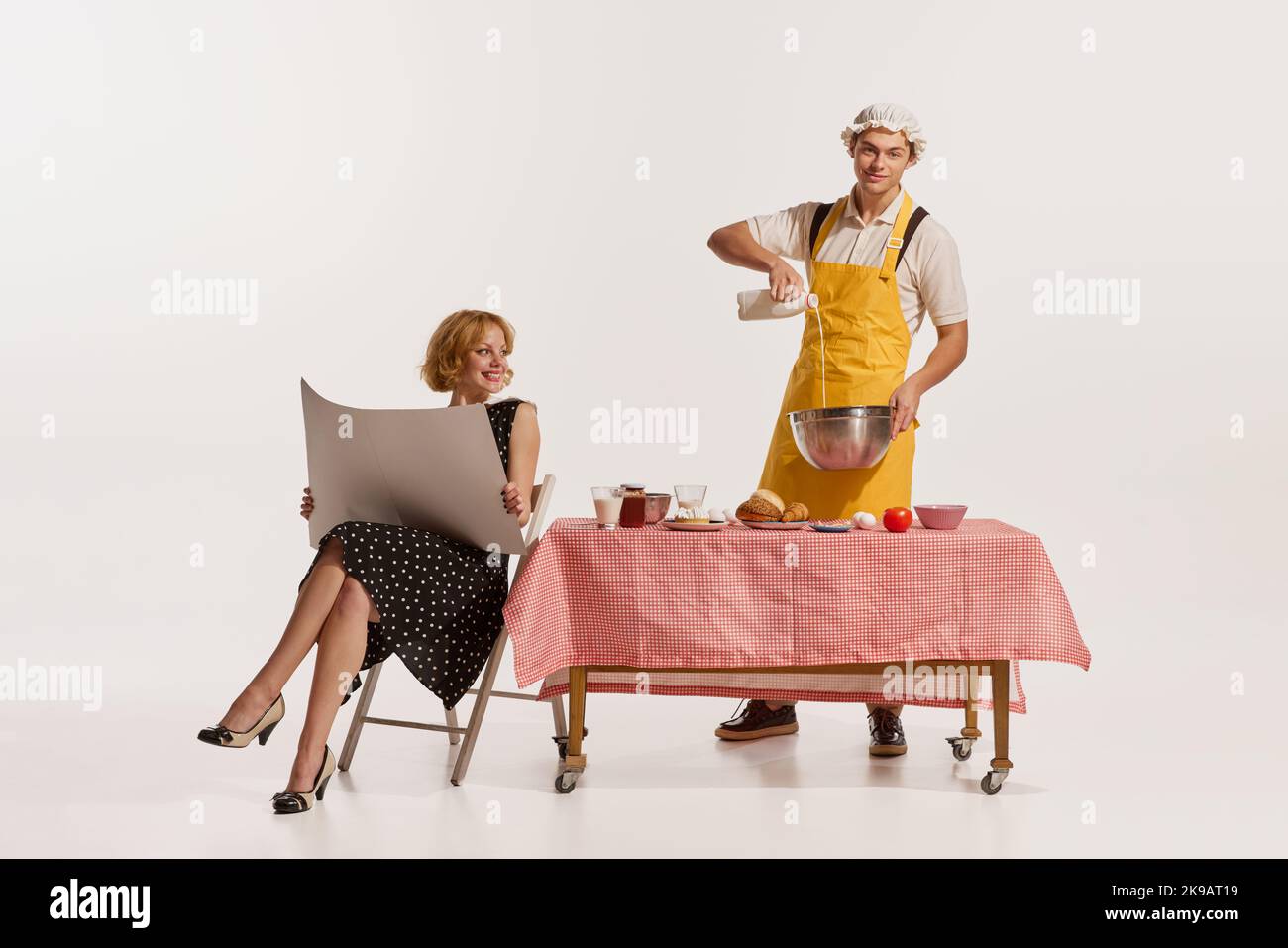 Porträt eines fröhlichen Mannes in Schürze Kochen, während Frau am Tisch sitzen und Zeitung isoliert auf weißem Hintergrund lesen Stockfoto