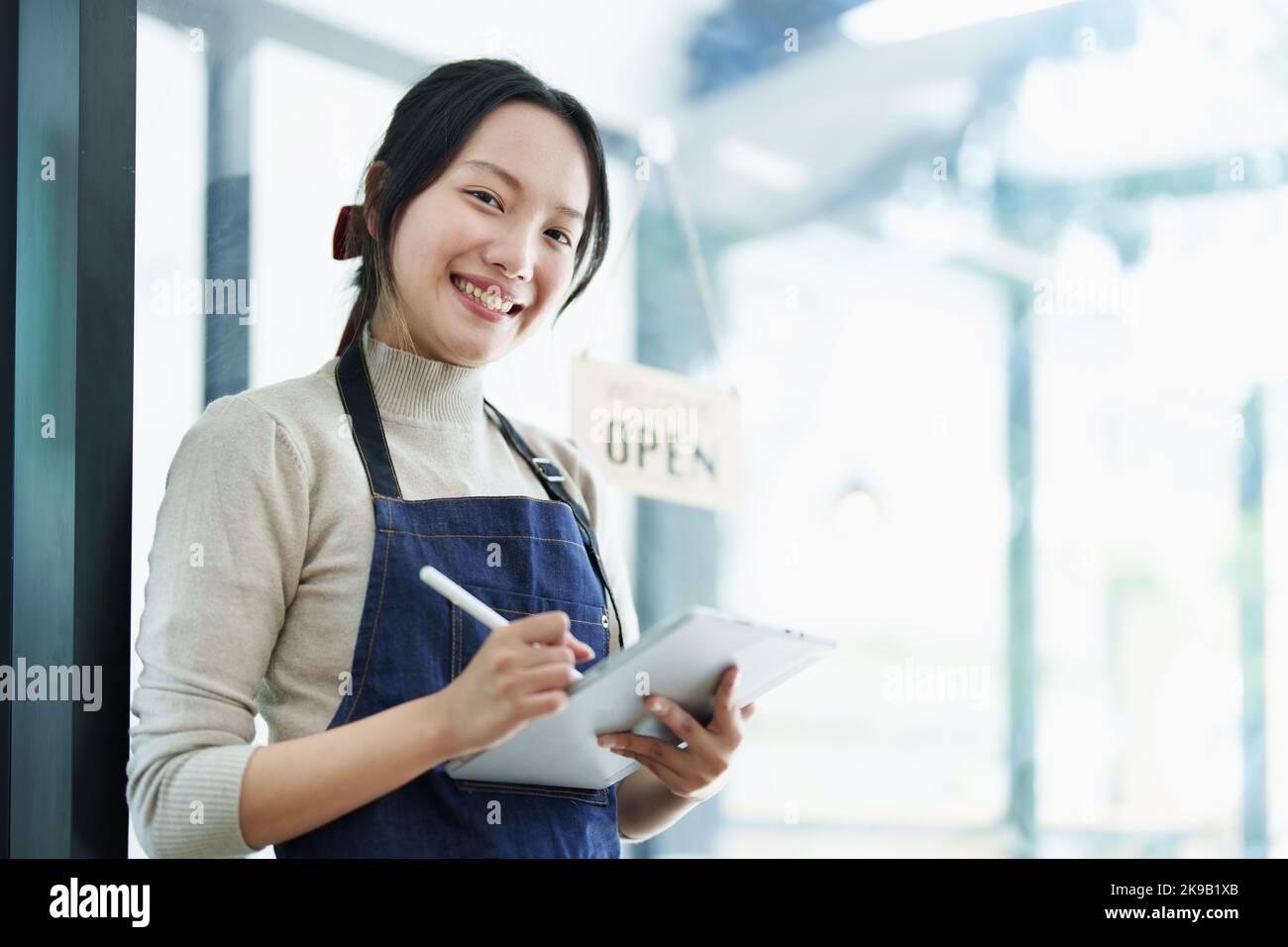 Eine junge asiatische Frau, die ein kleines Unternehmen eröffnet und ein lächelndes Gesicht zeigt, hält ein Tablet in einer Schürze, die vor einer Café-Bar steht Stockfoto