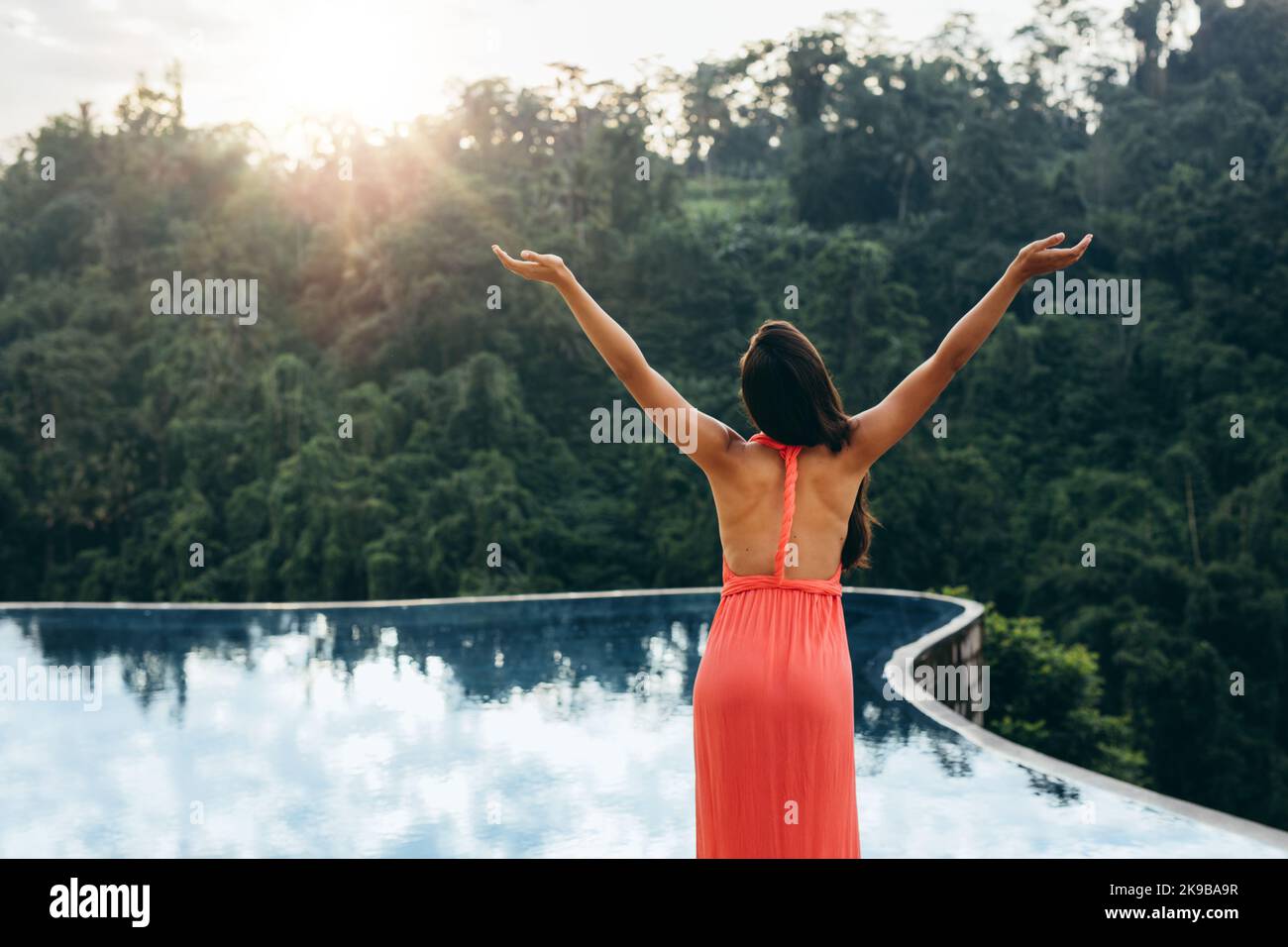 Rückansicht Porträt einer schönen jungen Frau, die mit erhobenen Händen am Pool steht und in den Himmel blickt. Weibliches Modell, das tropische Klippe genießt Stockfoto