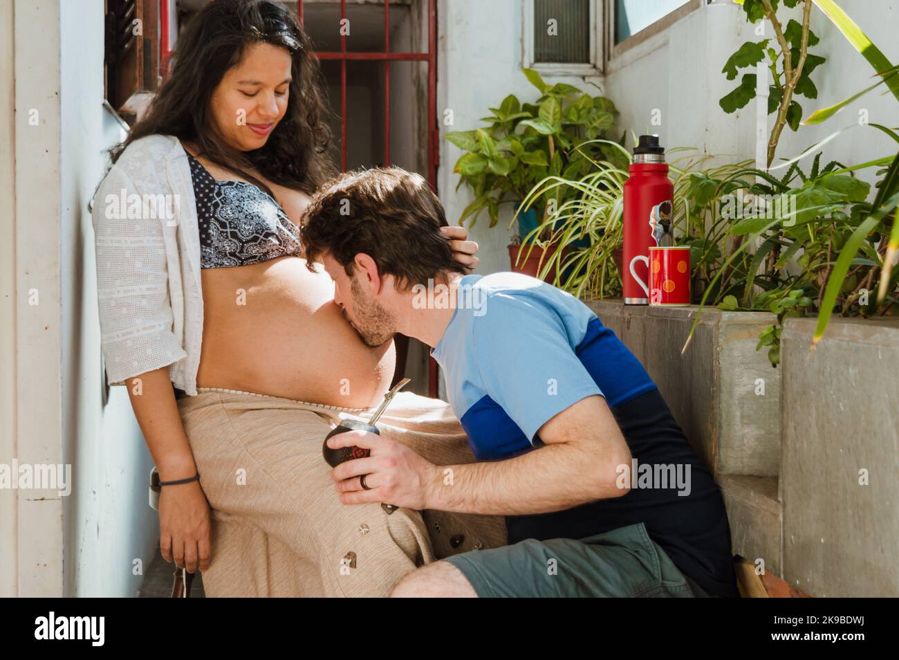 Der junge lateinische Kaukasusmann zu Hause küsst den Bauch seiner Frau, die schwanger ist, und sitzt glücklich und sieht zu, wie er seinem nächsten Baby Liebe schenkt. Stockfoto
