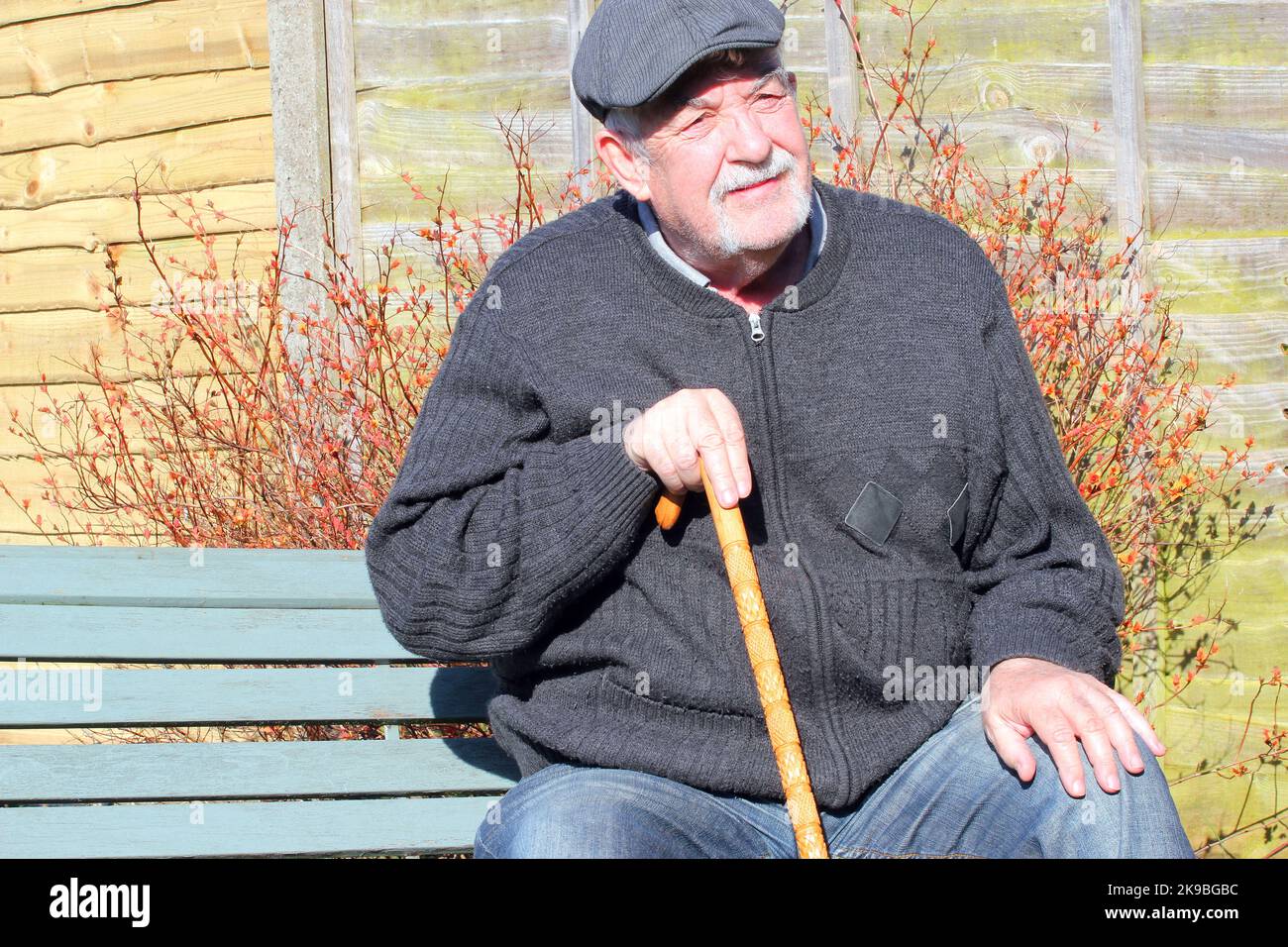 Alter oder älterer Mann, der auf einer Bank sitzt und sich entspannt. Mann, der eine Stoffkappe trägt und einen Spazierstock hält. Stockfoto