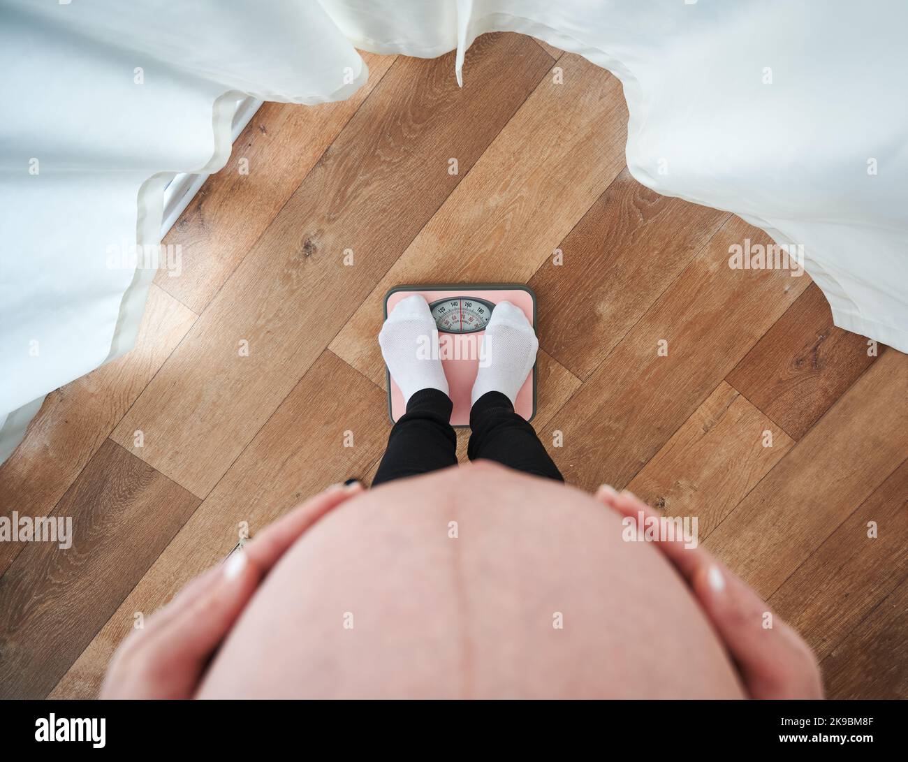 Draufsicht auf die werdende Mutter, die zum Wiegen auf der Bodenwaage steht und ihre beiden Handflächen auf den Bauch legt. Eine Schwangerin misst ihr Körpergewicht zu Hause mit einer Personenwaage. Stockfoto