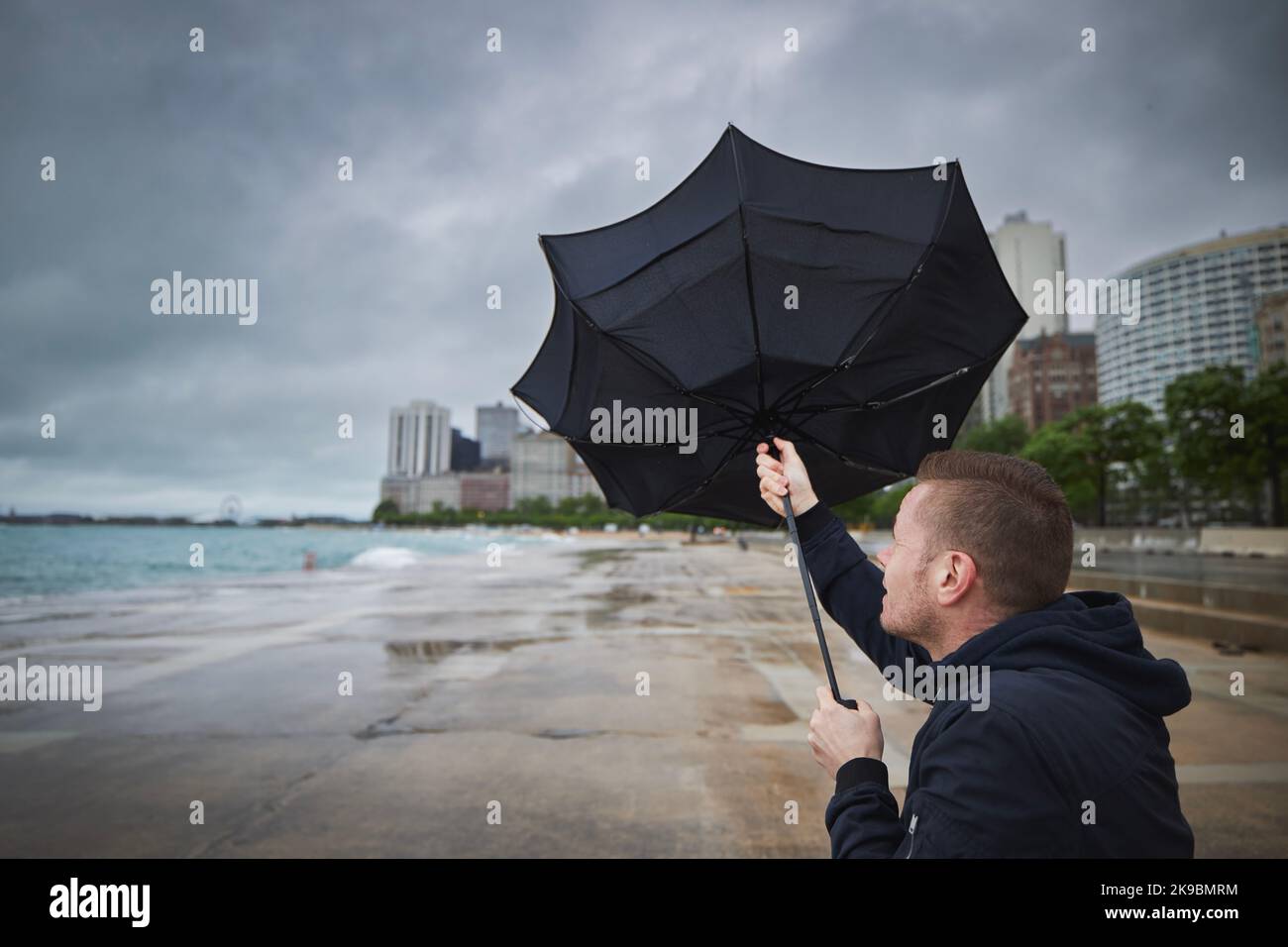 Mann hält gebrochenen Regenschirm bei starkem Wind während düsteren regnerischen Tag in der Stadt. Stockfoto