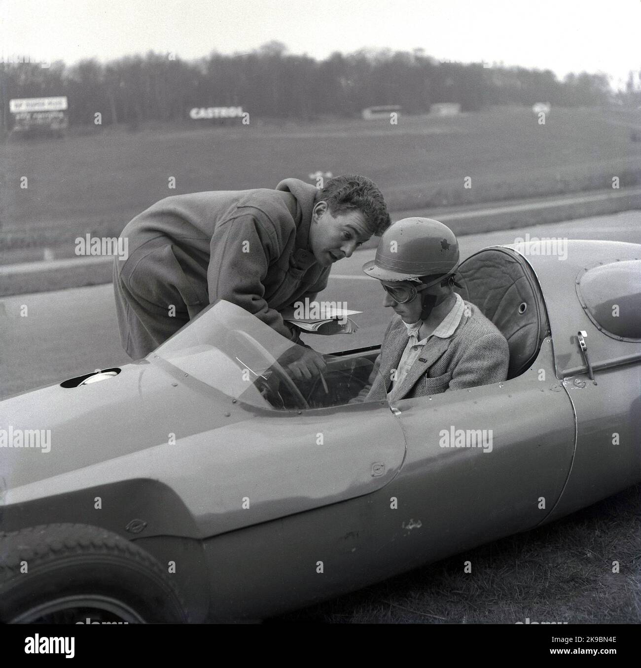 1959, historisch, Motorsport, Rennsport Fahrschule. Das Bild zeigt einen männlichen Instruktor, der mit einem Fahrer spricht, der in einem Cooper-Auto auf der Rennstrecke auf der Rennstrecke von Brands Hatch in Kent, England, Großbritannien, sitzt. Der Fahrer trägt eine Sportjacke und einen Helm und googles of the era. Zu dieser Zeit war die Coopers Driving School auf der Rennstrecke beheimatt. Stockfoto