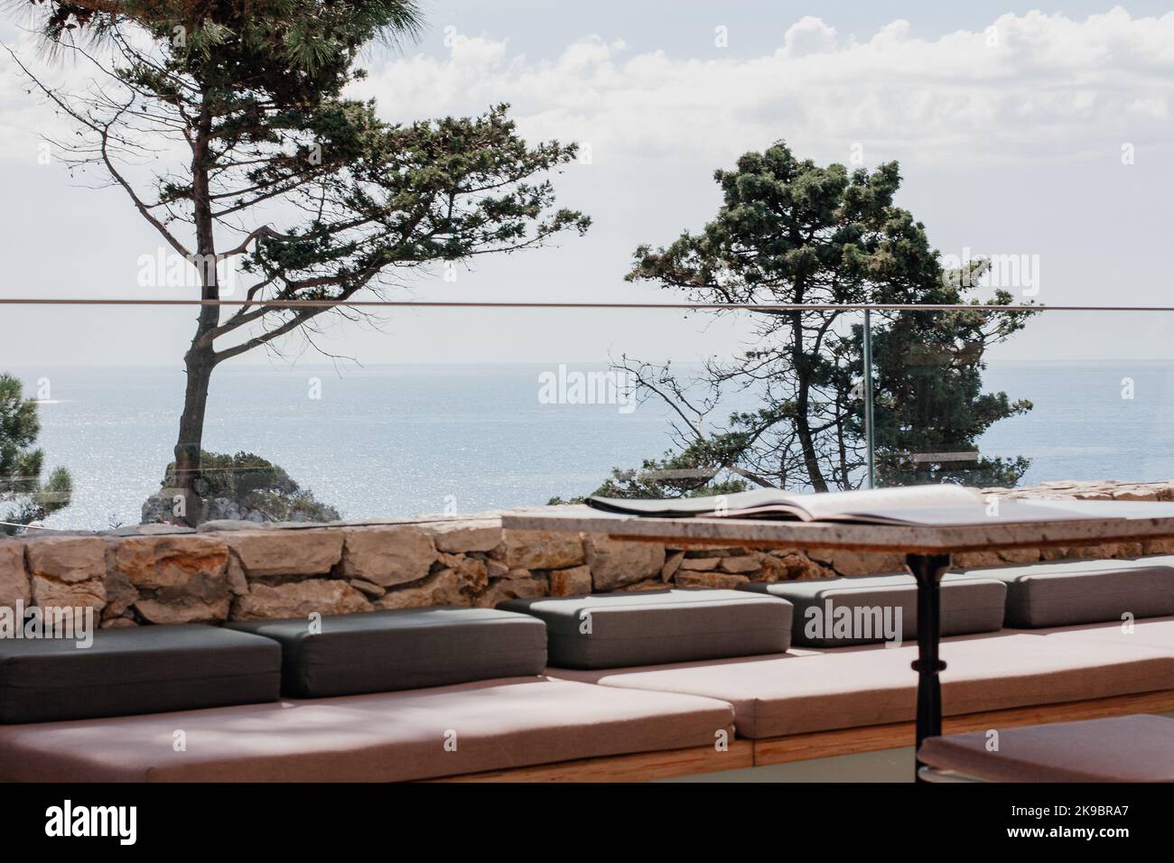 Korfu, Griechenland. Die Veranda des Cafés mit einer Glasgrenze mit Blick auf den Strand und blau im Wasser. Couchtisch und weiche Kissen zum Sitzen. Stockfoto